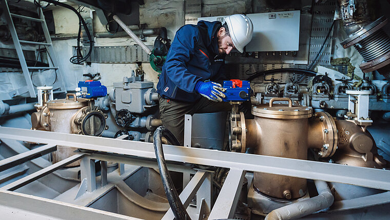 A person wearing blue protective clothing and a white helmet is working in a machine environment. In front of the person are large metal components, including two bronze-coloured valves and pipes. Further technical parts, cables and fastenings are visible in the background.