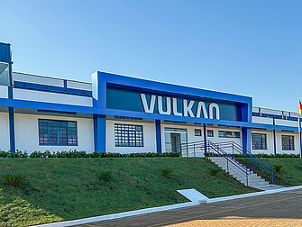 Wide exterior view of a white and blue industrial or commercial building with large lettering reading “VULKAN” above the entrance; in front of it are green spaces, a staircase, and a clear blue sky.