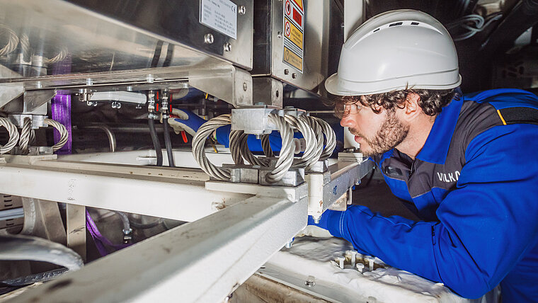 Uma pessoa veste roupa de proteção azul e um capacete branco e trabalha em uma tubulação metálica em um ambiente industrial. Ao redor da tubulação, são visíveis várias válvulas, mangueiras e fixações. Ao fundo, há outros tubos, cabos e componentes técnicos.