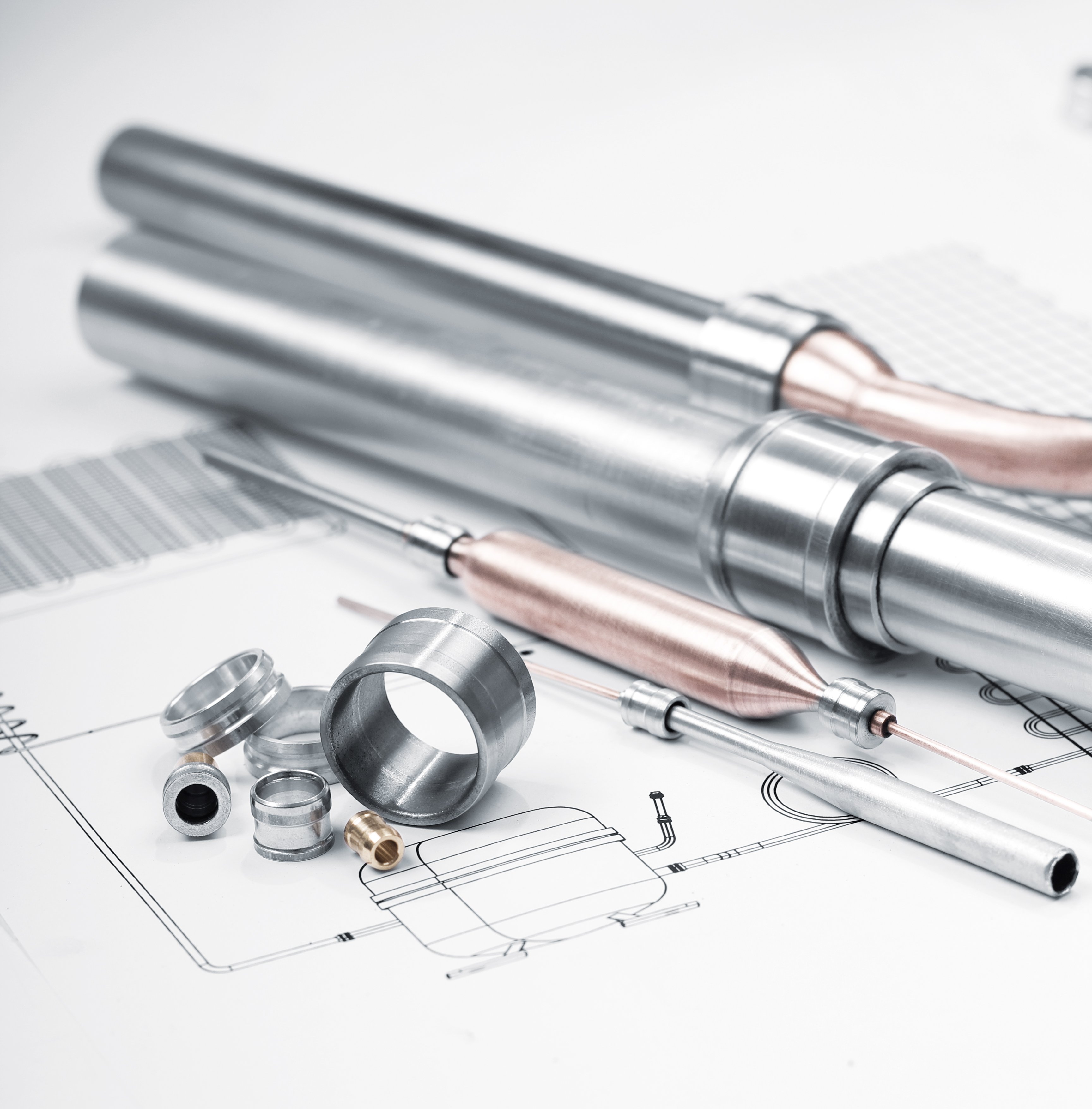 Zwei große Metallrohre, einige kleinere Metallverbindungen, ein Bleistift und ein Lineal liegen auf technischen Zeichnungen oder Bauplänen. Die Zeichnungen zeigen verschiedene Linien und Maßangaben.