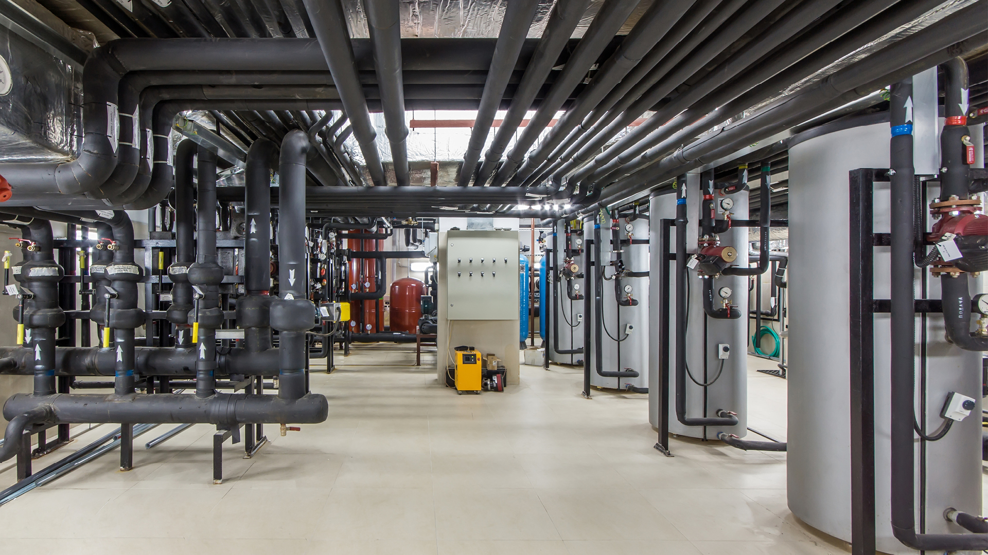 Sala técnica con equipos industriales como tuberías, tanques y elementos de control, bien iluminada y ordenada, con tuberías negras en el techo y las paredes, así como válvulas e indicadores de colores.