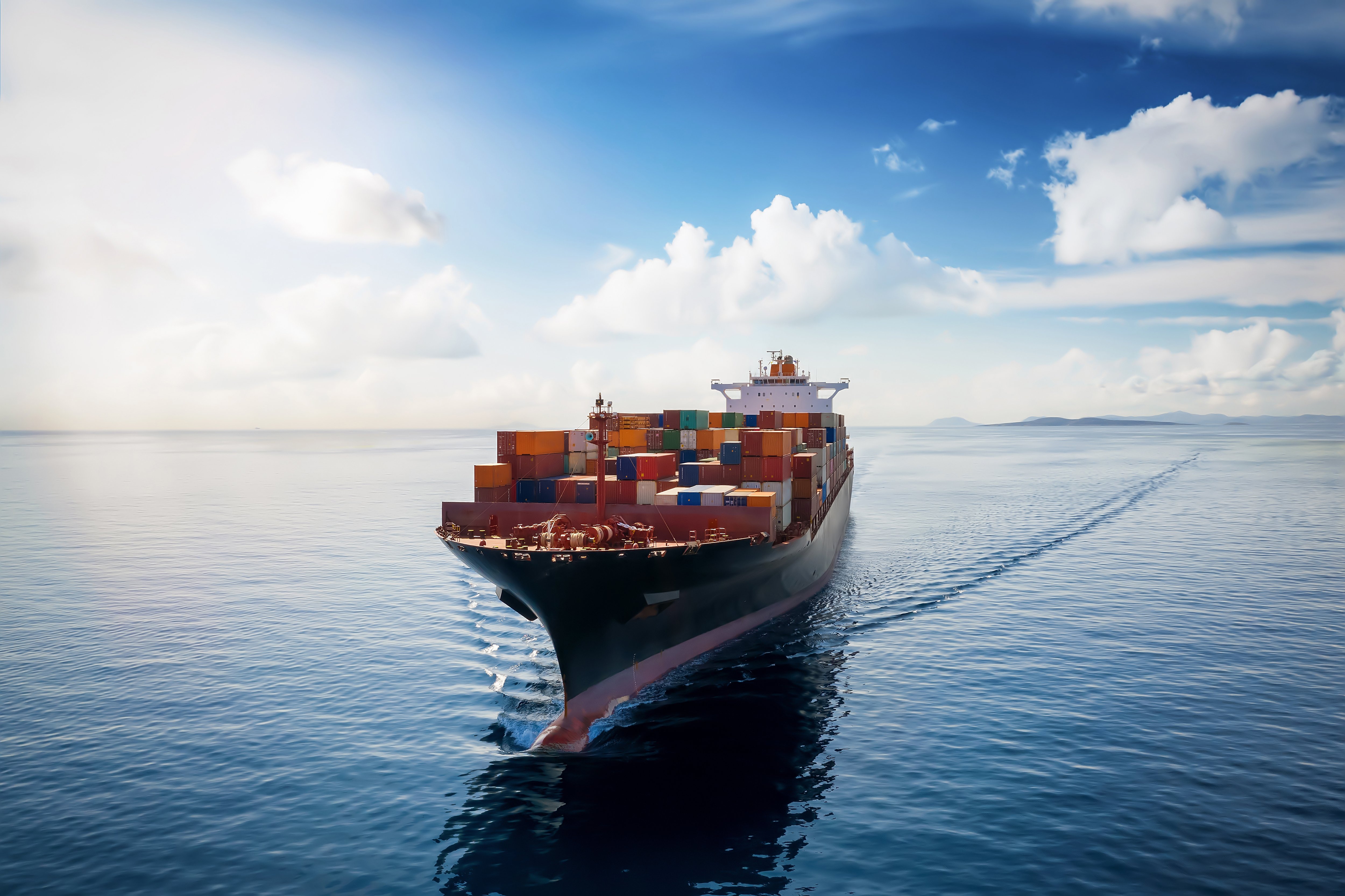 Ein großes Frachtschiff fährt auf dem Ozean. Auf dem Deck sind zahlreiche Container gestapelt. Der Himmel ist teilweise bewölkt, und das Wasser zeigt sanfte Wellen.