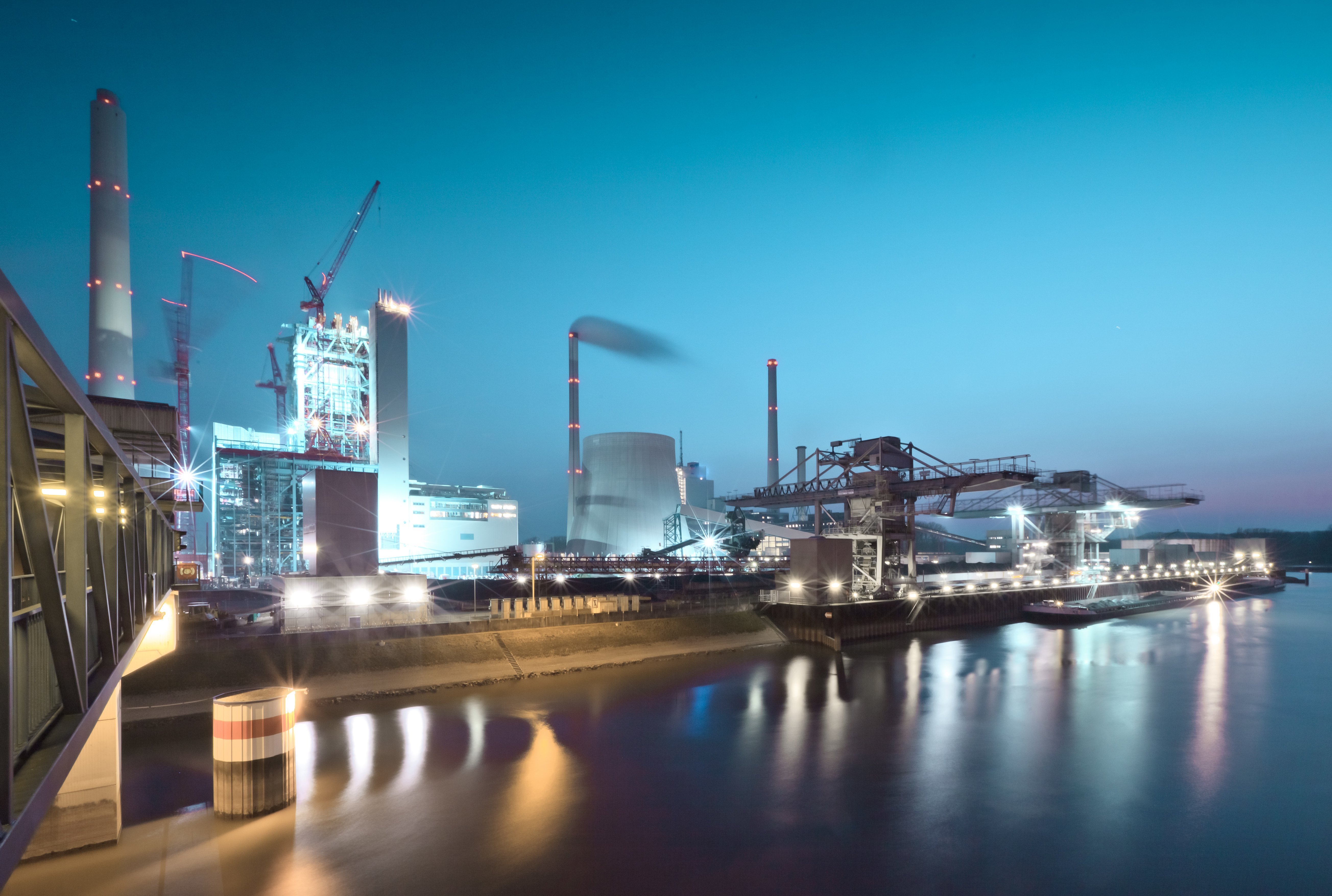 Área industrial ao entardecer com prédios iluminados, guindastes e chaminés. Reflexos visíveis nas águas adjacentes.