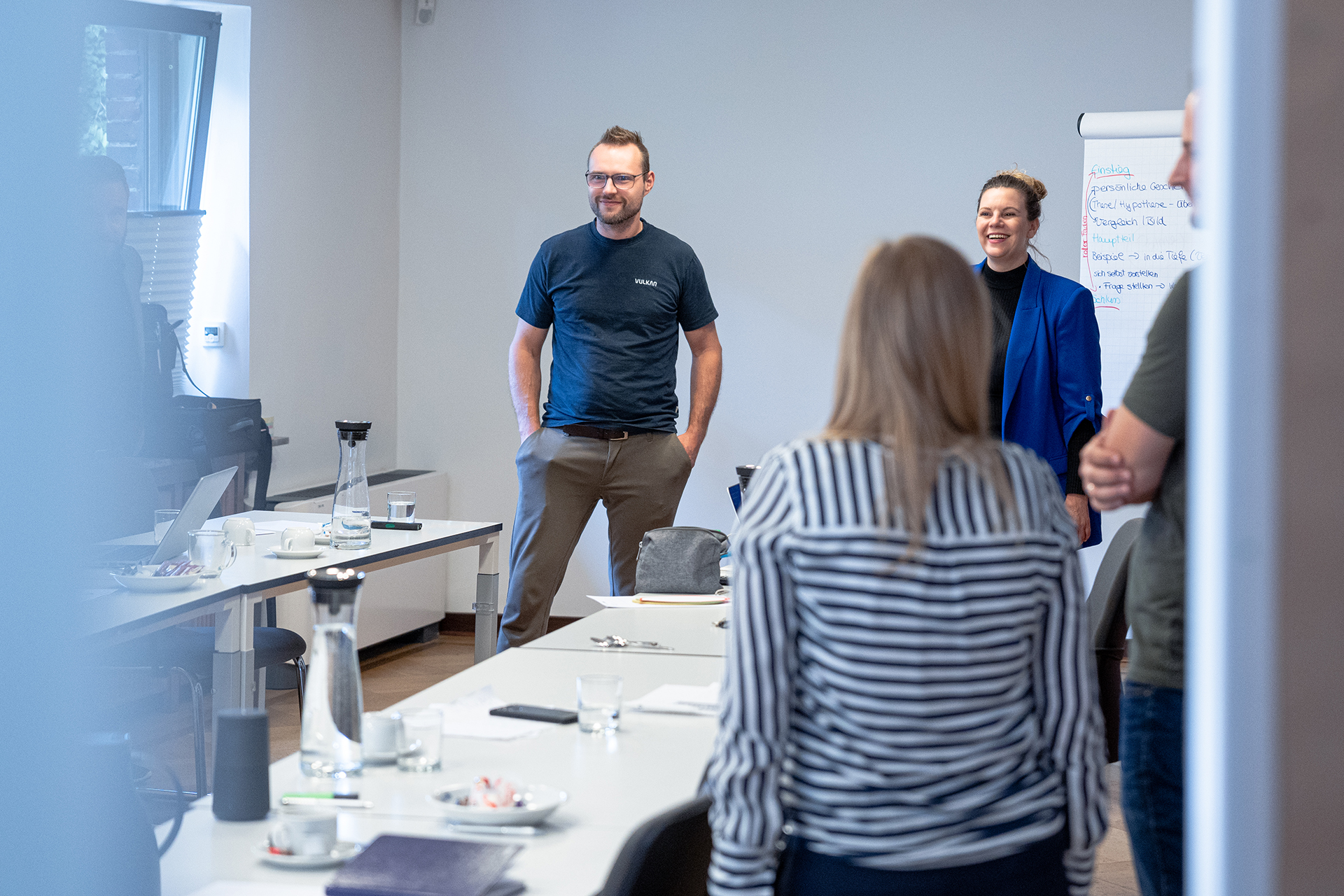 A group of four people (3 men, 1 woman) are standing in a meeting room. One person has their back to the camera, three people are facing the camera. Various items are on the table, including paper, a laptop, and a coffee mug. There is a whiteboard or flipchart in the background.