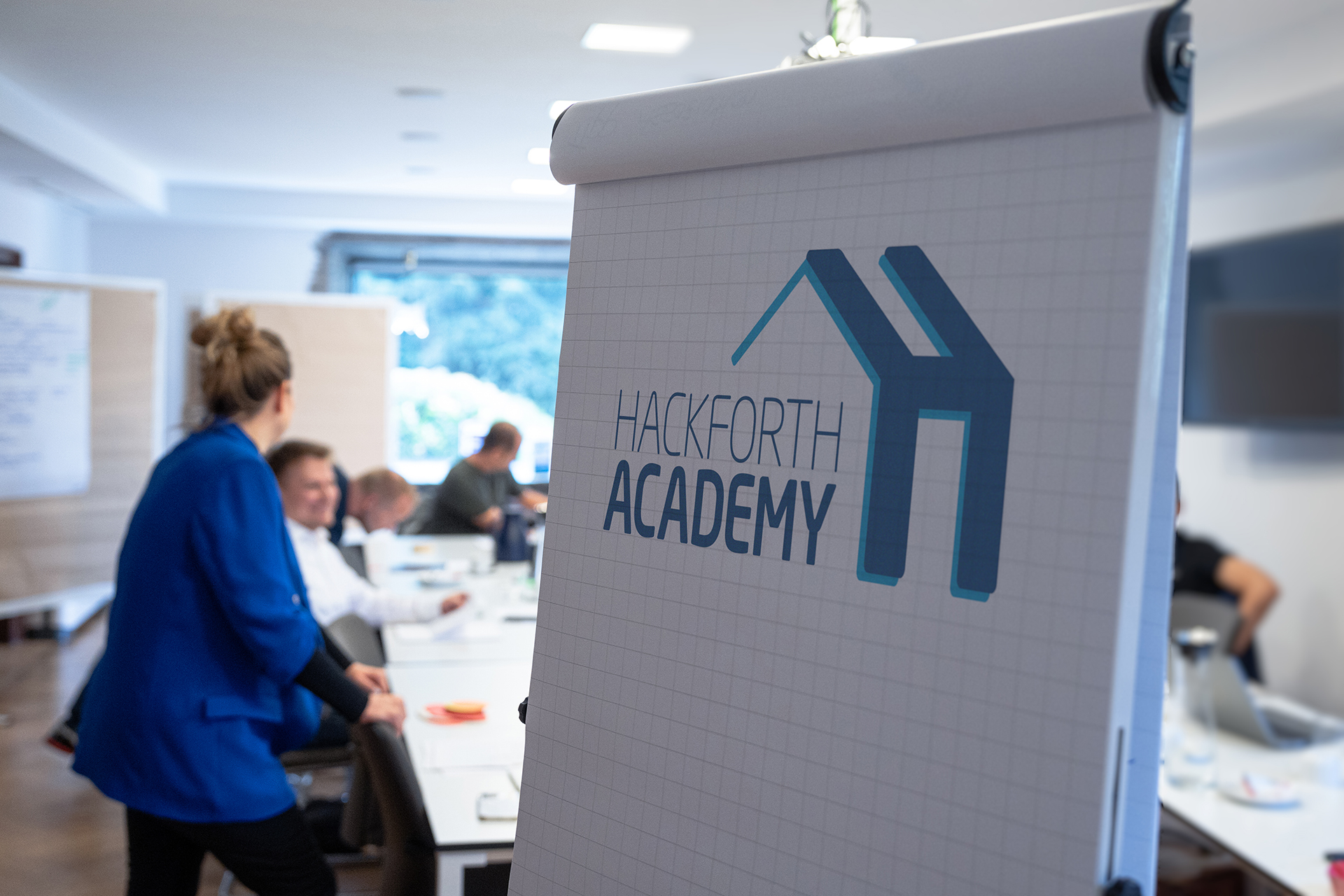 A group of people are in a meeting room. In the foreground is a flipchart displaying the Hackforth Academy logo.