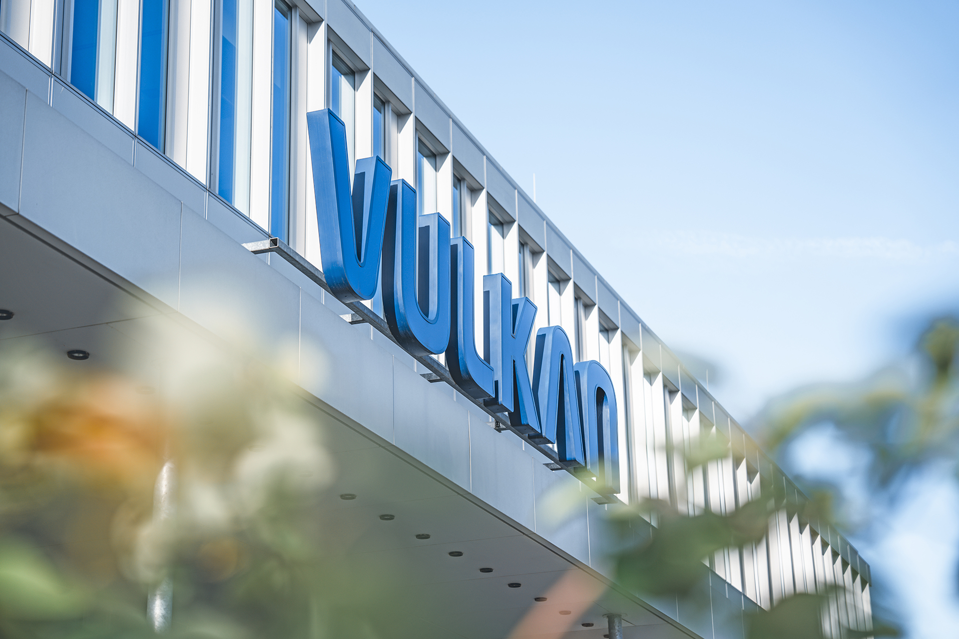 Oblique exterior view of a building with large blue lettering “VULKAN” on the facade; blurred plants in the foreground.