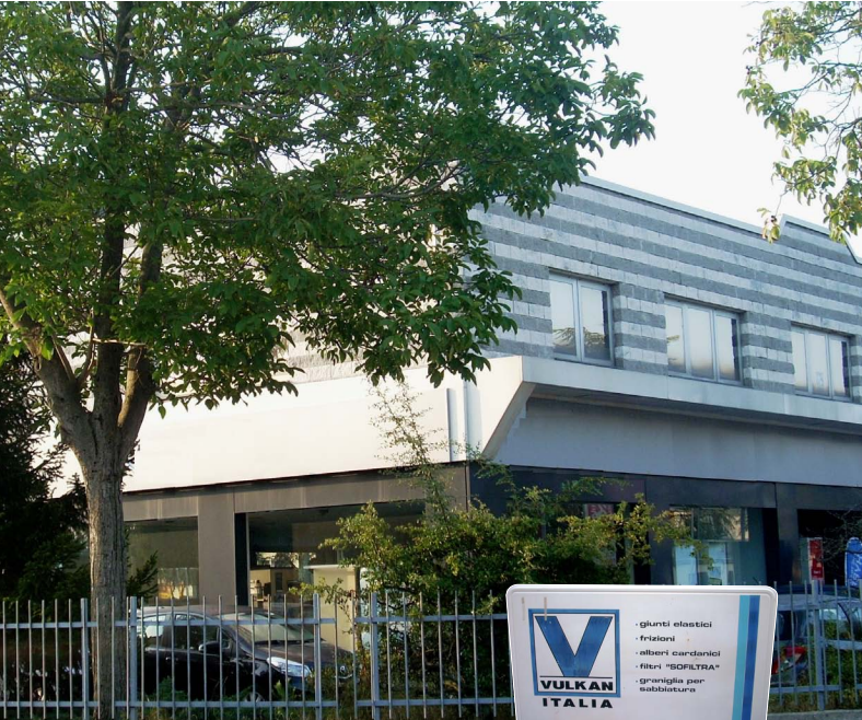 Modernes Firmengebäude mit grauer Fassade und großen Fenstern, teilweise von Bäumen verdeckt. Im Vordergrund steht ein weißes Schild mit blauem Logo und der Aufschrift ‚VULKAN‘. Ein Metallzaun verläuft entlang der Vorderseite des Geländes.