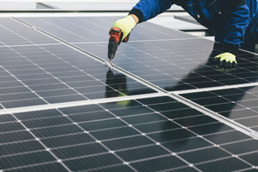 Uma pessoa trabalha com uma chave de fenda elétrica na superfície de painéis solares. A pessoa usa luvas amarelas e roupas azuis.