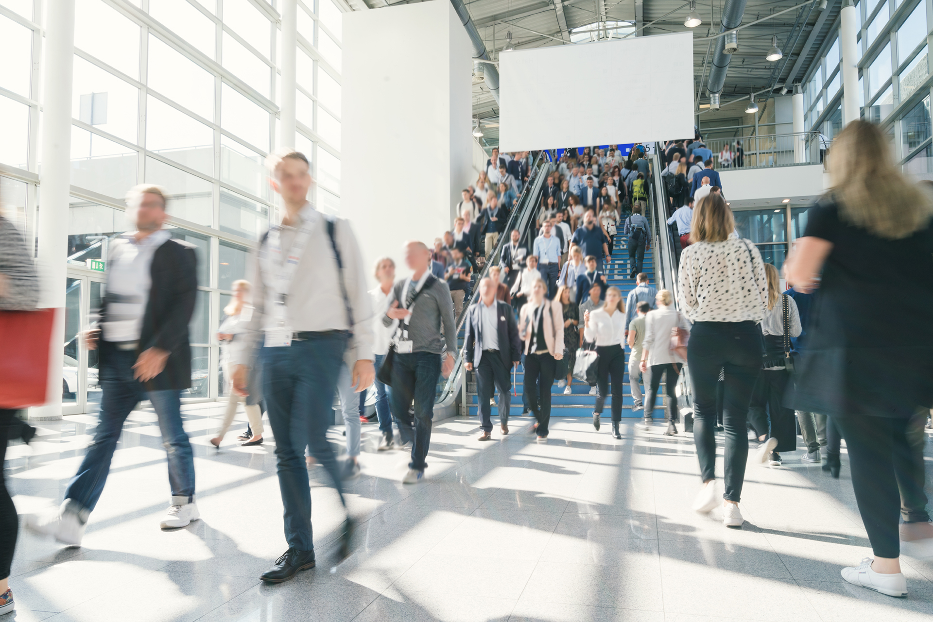 Many people walk through a large, bright hall with glass walls and a wide staircase. Some people climb the stairs, others move around on the floor area.